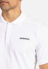 Valkoinen polo-paita, joka on valmistettu teksturoidusta kankaasta. Paidassa on perinteinen kaulus ja kolmen napin napitus. Musta "ROSSIGNOL" -logot vasemmalla puolella.