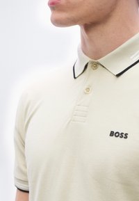 Homme portant un polo beige avec un bord noir sur le col et les manches, arborant le logo « BOSS » sur le côté gauche de la poitrine.