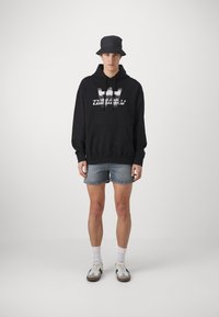 Zwarte hoodie met voorzak en grafische logo, gecombineerd met lichtblauwe denim shorts en een zwarte bucket hat, met casual sneakers.
