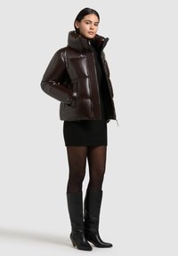 Brown, glossy puffer jacket with high collar, zip closure, and side pockets. Worn over a black sweater dress and paired with black knee-high boots.