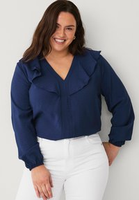 Blusa a maniche lunghe blu navy con dettagli arricciati lungo il collo, tessuto testurizzato, scollatura a V e polsini elasticizzati, abbinata a pantaloni bianchi.