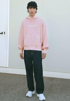 Giovane con capelli ricci che indossa un hoodie rosa con la scritta "SILENT ICONS", jeans scuri e sneakers bianche, in piedi su un marciapiede all'aperto.