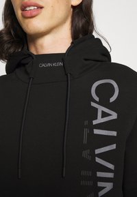 Černá mikina s vysokým krkem, která má šňůrku na stažení. Logo "CALVIN KLEIN" v šedém vertikálním tisku na pravé straně. Jemná textura látky.