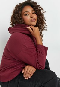 Felpa bordeaux con cappuccio dal finish morbido e testurizzato. Presenta una vestibilità comoda e maniche ampie. Il modello è seduto comodamente con le mani sulle ginocchia.