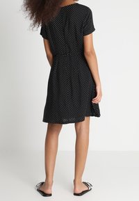 Robe noire à pois avec des manches courtes, taille cintrée et jupe fluide. Associée à des sandales plates avec un accent de nœud décoratif.