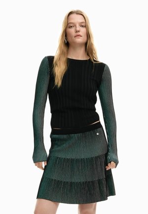 Žena v čiernom rebrovanom tričku s dlhými prúžkovanými zeleno-čiernymi rukávmi, doplnená o zodpovedajúcu plisovanú sukňu.