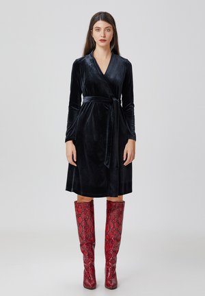 Robe enveloppante en velours noir avec des manches longues et une taille à nouer, accompagnée de bottes rouges à motif serpent montantes jusqu'aux genoux. Texture lisse et forme structurée.