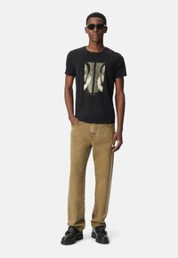 Uomo che indossa una t-shirt grafica nera, jeans beige, scarpe nere e occhiali da sole scuri, in piedi con una mano in tasca su uno sfondo semplice.