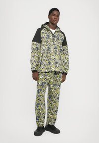 Fargerik trykt joggedress med et blomsterdesign i grønt, blått og hvitt, sammen med en svart hettejakke og matchende bukser.
