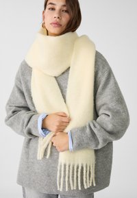 Sciarpa di lana crema con orli frangiati, indossata in modo morbido sopra un maglione grigio chiaro. Finitura morbida e texture, abbinata a una camicia blu con colletto sotto.
