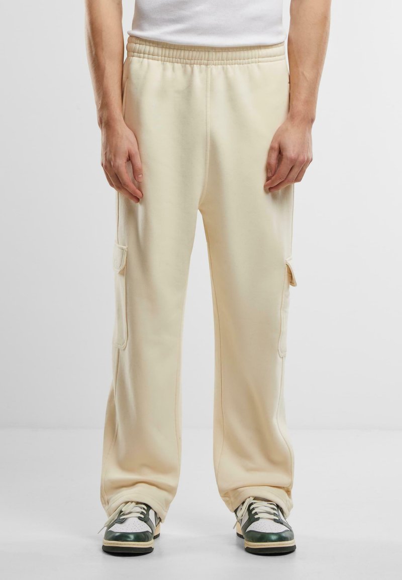 Pantalons de jogging en coton beige avec poches cargo latérales, taille élastique et coupe décontractée, assortis à des baskets vertes et blanches.