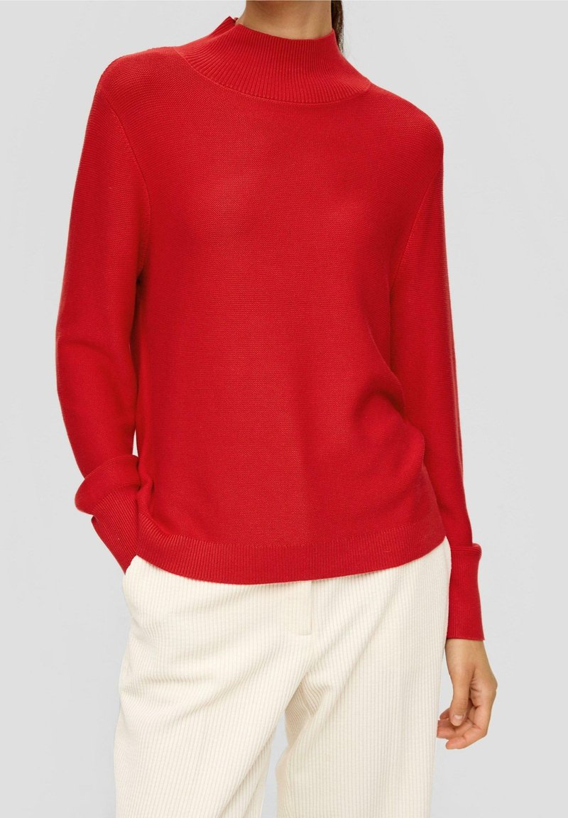 Roter Strickpullover mit hohem Ripp-Ausschnitt und langen Ärmeln, in lockerer Passform. Getragen mit cremefarbenen gerippten Hosen.