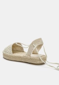 Sandale espadrille en tissu tissé beige, avec une semelle en jute tressé et des sangles de cheville croisées. Design ouvert avec des détails texturés.