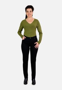 Camiseta verde de manga larga con escote en V y detalles de botones, combinada con jeans negros ajustados y botas negras de tobillo, de pie sobre un fondo liso.