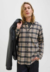 Beige und schwarzes Karobluse aus Flanellmaterial, mit einem Knopfverschluss und einer lockeren Passform, getragen mit einer schwarzen Lederjacke, die über der Schulter hängt.