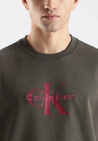 Sudadera de color verde oliva con cuello redondo, que presenta un bordado en relieve del logo "Calvin Klein" en burdeos en el centro. Textura de tela suave.