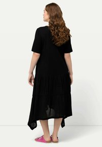 Schwarzes, gestuftes Kleid aus leichtem Stoff, mit rundem Ausschnitt und kurzen Ärmeln. Akzentuiert mit einem asymmetrischen Saum. Tragen Sie es mit rosa Sandalen.