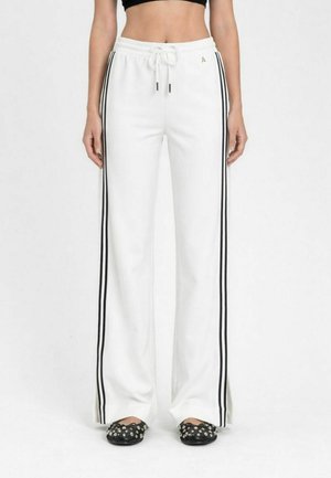 Donna che indossa pantaloni bianchi a gamba larga con strisce nere laterali, vita con coulisse e scarpe slip-on nere con borchie.