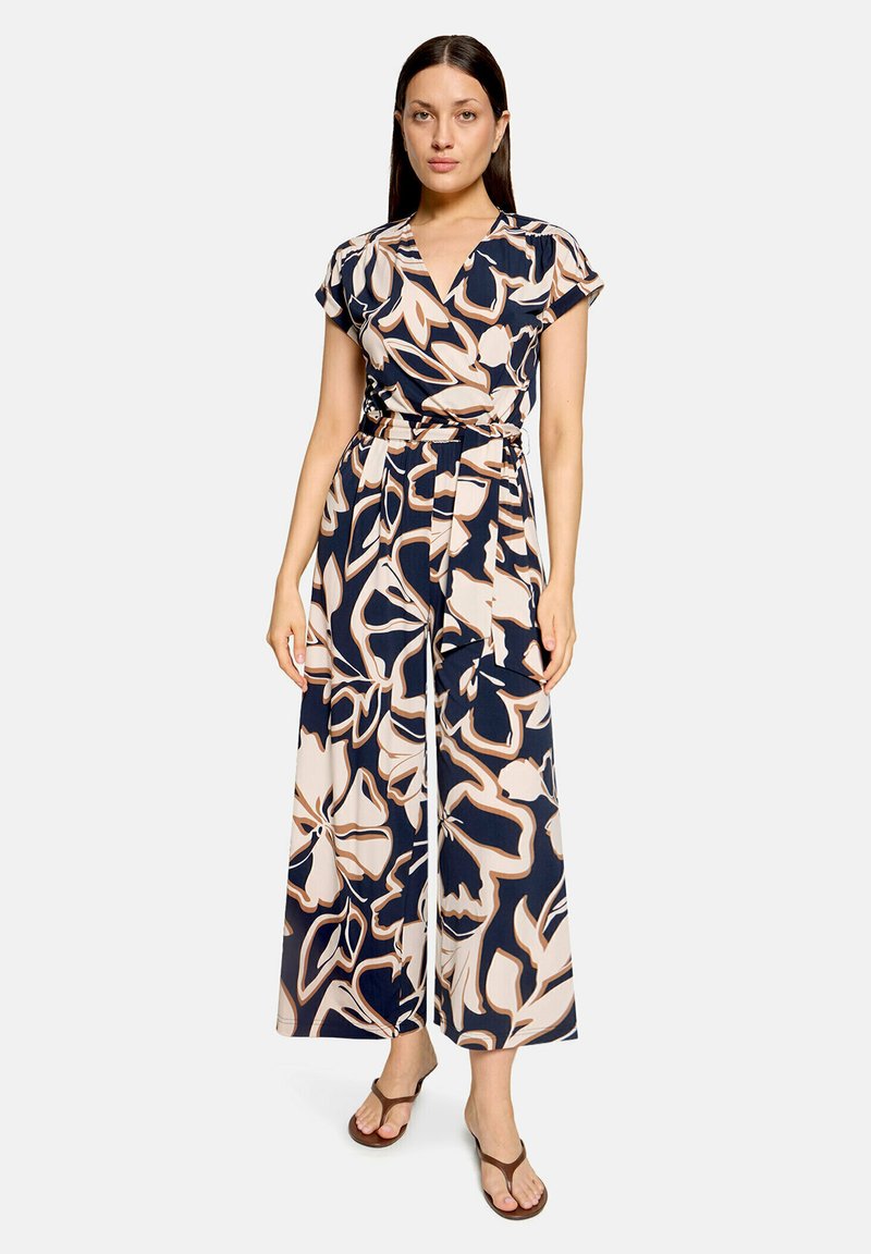 Femme portant une combinaison à manches courtes ceinturée avec des jambes larges présentant un grand motif floral abstrait beige et bleu marine et des sandales marron.
