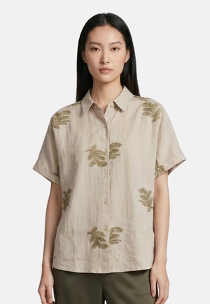 Giovane donna con lunghi capelli neri che indossa una camicia beige a maniche corte con bottoni e motivi di foglie marroni e pantaloni scuri.
