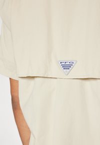 Camicia beige a maniche corte con orlo curvo, dotata di un logo PFG Performance Fishing Gear blu e argento sul retro. Design leggero e casual.