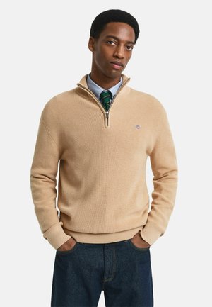 Uomo che indossa un pullover beige a zip quarti sopra una camicia blu con una cravatta a righe verdi, mani nelle tasche, in piedi contro uno sfondo bianco.