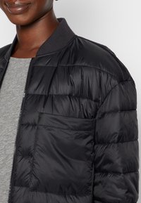 Chaqueta de abrigo negra con un acabado texturizado, que presenta un cuello acanalado y un cierre de cremallera. Incluye un bolsillo cuadrado en la parte frontal.