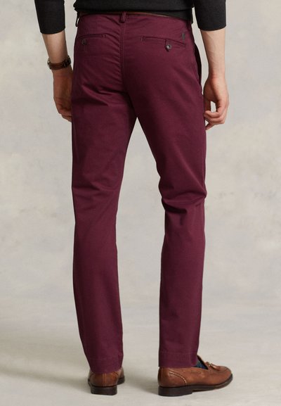 Άνδρας που φοράει μπορντό παντελόνια slim-fit, καφέ δερμάτινα παπούτσια και μαύρο πουκάμισο με μακριά μανίκια, στέκεται με τα χέρια χαλαρά στο πλάι.