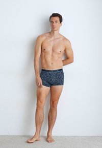 Kék mintás boxeralsó fekete derékrésszel, geometrikus mintázattal. A modell mezítláb áll egy sima világos fal előtt.