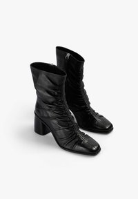 Botines de cuero negro con diseño fruncido, puntera cuadrada y tacón grueso. Cuentan con cremallera lateral para facilitar su uso.