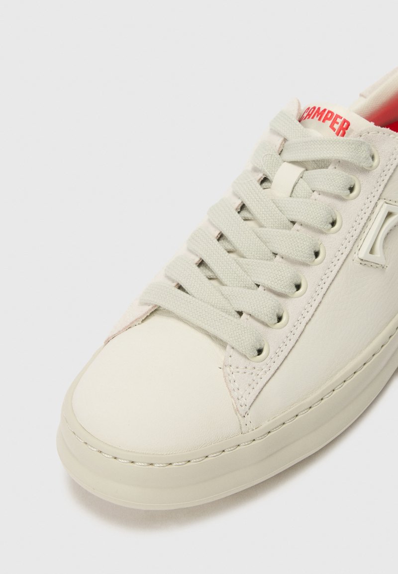 Weiße Leder-Sneaker mit flachen Schnürsenkeln, einer strukturierten Zunge und einem roten Logo an der Ferse. Abgerundete Zehenkappe mit genähten Akzenten entlang der Nähte.