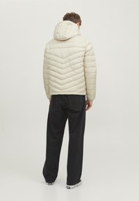 Giacca trapuntata beige chiaro con cappuccio, caratterizzata da un motivo a chevron; abbinata a jeans neri a gamba larga e sneakers bianche.