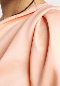 Hedvábně lososová saténová látka s jemnými sklady; hladký povrch s mírným leskem; detailní pohled na konstrukci oděvu.