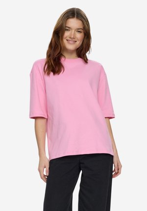 Kvinde med langt brunt hår iført en løs, lyserød kortærmet skjorte og sorte bukser, smilende med hænderne afslappet ved siden af kroppen.