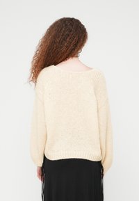 Γυναίκα με μακριά, σγουρά μαλλιά, που φοράει χαλαρό κρεμ πλεκτό πουλόβερ και μαύρη φούστα, όρθια με την πλάτη γυρισμένη, μπροστά σε απλό φόντο.