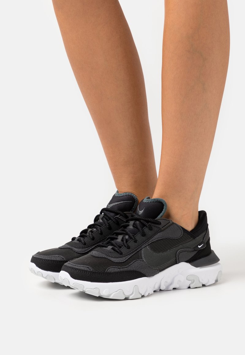 Zapatillas deportivas negras con una combinación de malla y materiales sintéticos, que presentan una suela blanca ondulada y sutiles acentos de logotipo.