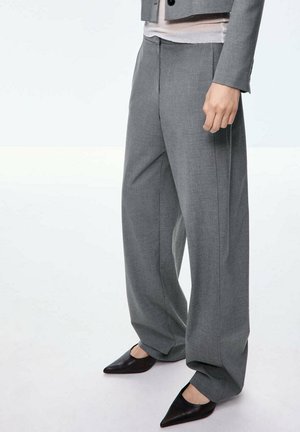 Persona que lleva pantalones grises sueltos, zapatos negros de punta y un blazer gris con una camisa blanca debajo.
