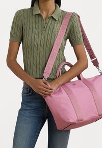 Rosa tote-väska med en slät yta, dubbla handtag och ett brett axelband med varumärkesnamnet. Stickad olivgrön polo.