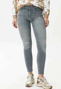 Jeans ajustados en denim gris deslavado con detalles desgastados. Combinados con zapatillas en tonos marrones y grises y un suéter de camuflaje beige.