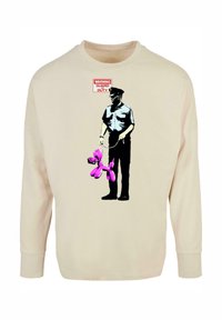 Beige Sweatshirt mit einem grafischen Motiv eines Polizeibeamten, der einen rosa Ballonhund hält, sowie einem Schild "ACHTUNG WACHDIENST". Lange Ärmel, lockere Passform.