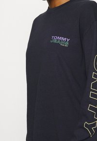 Marinblå långärmad T-shirt i bomull. Har "TOMMY JEANS"-logotypen i lila och grönt på bröstet, med ytterligare text i gult längs ärmen.