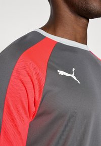 Puma sportpóló, szürke testtel, piros hálós vállakkal, fehér logóval és világosszürke kivágással. Simára szőtt anyag, eltérő textúrákkal.