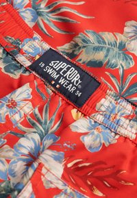Bañador rojo con un vibrante patrón floral en azul y blanco. Cintura elástica con una etiqueta que dice "Superdry 19 SWIM WEAR."