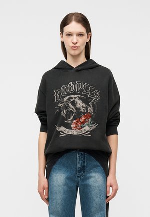 Νεαρή γυναίκα που φοράει μαύρο φούτερ με κουκούλα, διακοσμημένο με σχέδιο λιοντάρι πανθηρα και κόκκινα λουλούδια, μπλε τζιν με δύο αποχρώσεις, στέκεται μπροστά σε απλό φόντο.