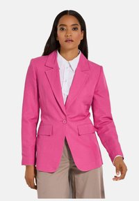 Aansluitende roze blazer met een enkele knoop, notch revers en twee voorzakken. Geweven stof, getailleerd silhouet, gedragen over een wit shirt.