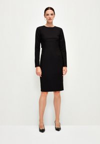 Abito lungo nero a maniche lunghe, attillato, con scollatura tonda, fino al ginocchio, realizzato in tessuto liscio. Indossato con scarpe nere con dettagli dorati.