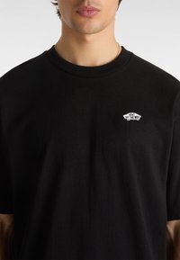 Camiseta de algodón negra con cuello redondo, que cuenta con un parche de logo blanco que dice "VANS OFF THE WALL" en la parte superior izquierda.