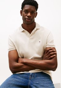 Béžové polo tričko s límečkem a krátkými rukávy, s malým vyšívaným logem. Kombinováno s modrými džíny, které zdůrazňují strukturu látky a střih.