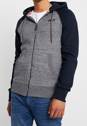 Homme portant un sweat à capuche gris zippé avec des manches et une capuche bleu marine, des poches avant et un jean bleu, debout devant un fond uni.
