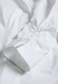 Blusa bianca con dettagli delle maniche arricciate e polsini strutturati. Tessuto liscio con linee pulite. Il tessuto appare fresco e leggero.
